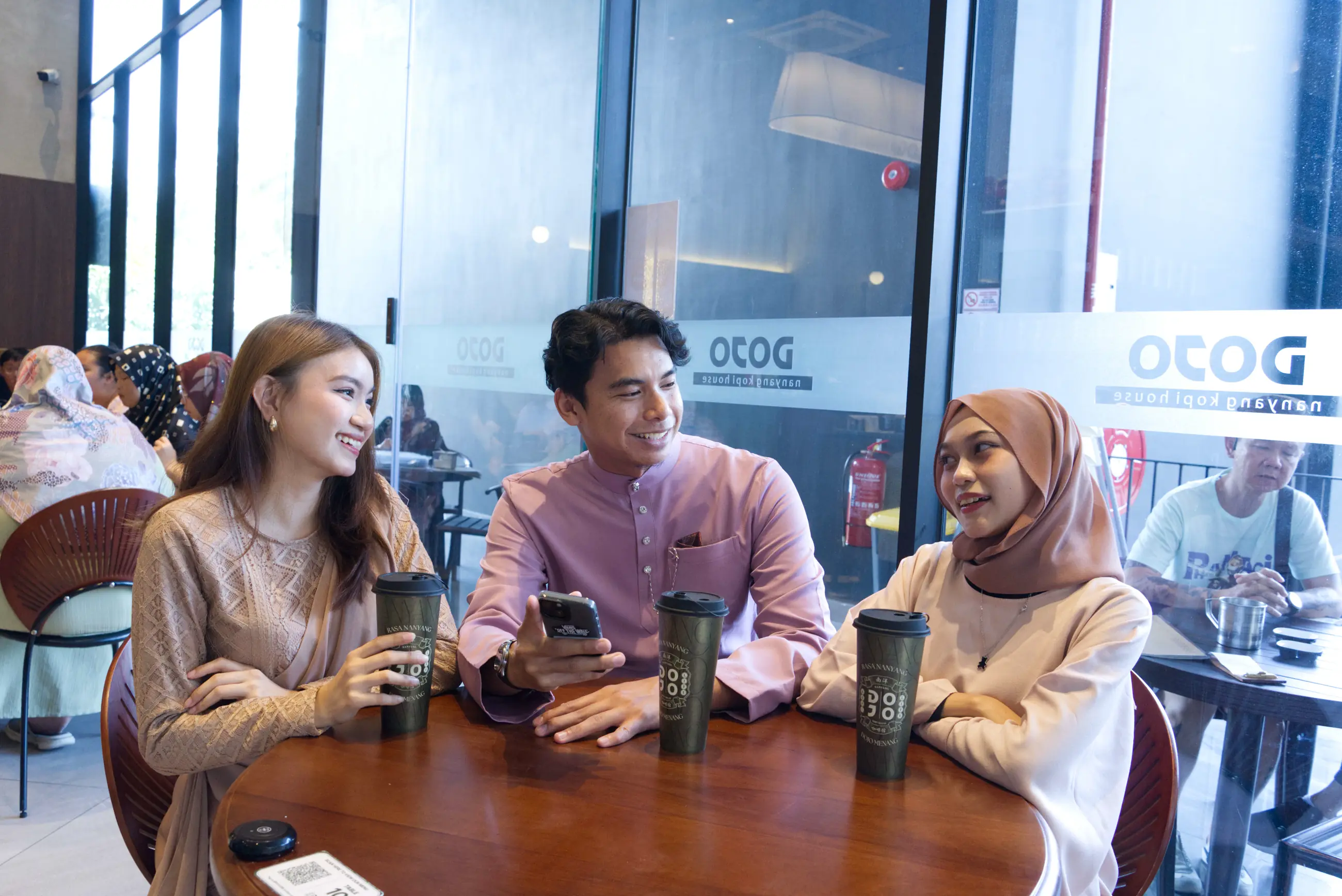 Three friends laughing over DOJO kopi at a table by the window